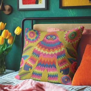 A square faux embroidery pillow featuring a brightly colored maximalist design against a mustard yellow background. In the center stands a stylized, furry Bigfoot or Sasquatch figure rendered in vibrant layers of pink, orange, blue, and lime green with a blue face and yellow eyes. The central creature is surrounded by large, detailed symmetrical pink and blue flowers and leaves. The flat printed fabric creates a highly realistic optical illusion of dimensional crewel embroidery stitches.
