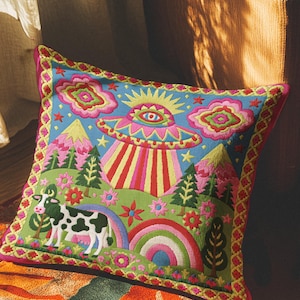 Description of a square throw pillow with a vibrant maximalist alien design. A central UFO with a large all-seeing eye hovers above a landscape with two rainbows, pink mountains, and green pine trees. A black and white spotted cow stands in the foreground. The design is a high-contrast palette of sky blue, magenta, hot pink, and meadow green. The pillow has a faux-embroidery 2D print finish on linen-feel fabric, framed by a yellow and pink scalloped floral border.