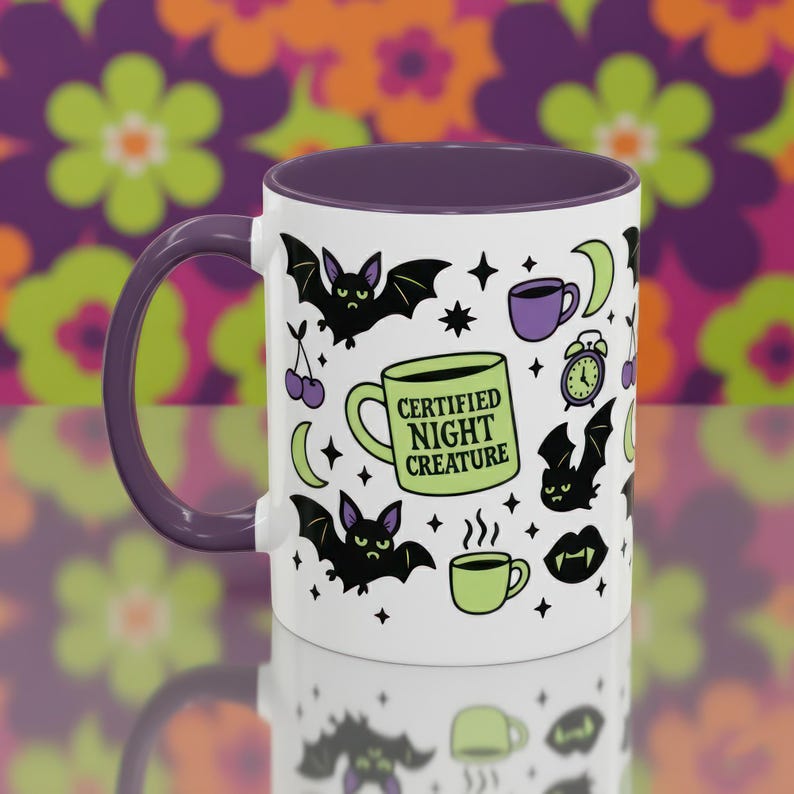 White ceramic mug with vibrant purple interior and matching C-handle. Features a full wrap-around, 'spooky cute' pastel goth design. Central graphic is a lime green coffee cup with 'CERTIFIED NIGHT CREATURE' in black text. Surrounding illustrations include grumpy black bats, a steaming green coffee cup, a purple mug, vampire fangs, crescent moon, black cherries, and an alarm clock. Design uses bold graphics in black, lime green, and purple, accented with small black stars. Pastel Goth vibes.