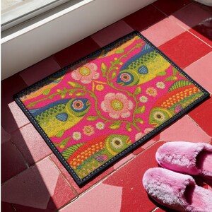 Rectangular indoor outdoor doormat featuring a vibrant hot pink background with a symmetrical maximalist pattern. The design showcases stylized sardines in yellow, lime green, and cobalt blue swimming horizontally towards a central vertical axis of blooming pale pink flowers and green vines. The imagery mimics the texture of vintage needlepoint embroidery but is a flat print on polyester. The mat is framed by a sleek black trim and features a non-slip rubber backing for safety.