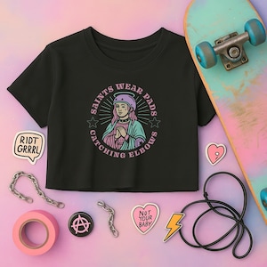 Puede incluir: Camiseta negra recortada con un gráfico de una mujer con casco y el texto "Saints Wear Pads Catching Elbows".