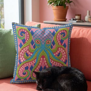 A square decorative throw pillow cover featuring a highly symmetrical bright octopus design. The octopus has a turquoise blue body with a diamond patterned head and eight curled tentacles detailed in bright pink and lime green. The background is a repeating diamond argyle pattern in royal blue and light pink. The artwork is a flat two dimensional print that mimics the textured look of intricate crewelwork embroidery. The pillow features a hidden zipper on the bottom edge. No insert included.