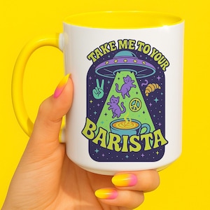 Puede incluir: Taza de cerámica blanca con interior y asa amarillos. La taza presenta un gráfico colorido de un OVNI abduciendo gatos morados, con el texto "TAKE ME TO YOUR BARISTA".