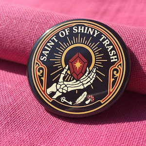 May include: A circular pinback button featuring a black background with a gold border. The design displays a skeletal hand holding a red gem, surrounded by the text SAINT OF SHINY TRASH, a small key, a feather, and a mushroom illustration.