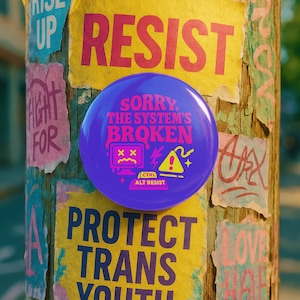May include: A circular purple pinback button featuring the text "SORRY, THE SYSTEM'S BROKEN" in pink, with a graphic of a computer monitor with "X" eyes, a yellow warning triangle, and a keyboard key labeled "CTRL ALT RESIST" in yellow.