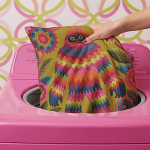 A square faux embroidery pillow featuring a brightly colored maximalist design against a mustard yellow background. In the center stands a stylized, furry Bigfoot or Sasquatch figure rendered in vibrant layers of pink, orange, blue, and lime green with a blue face and yellow eyes. The central creature is surrounded by large, detailed symmetrical pink and blue flowers and leaves. The flat printed fabric creates a highly realistic optical illusion of dimensional crewel embroidery stitches.
