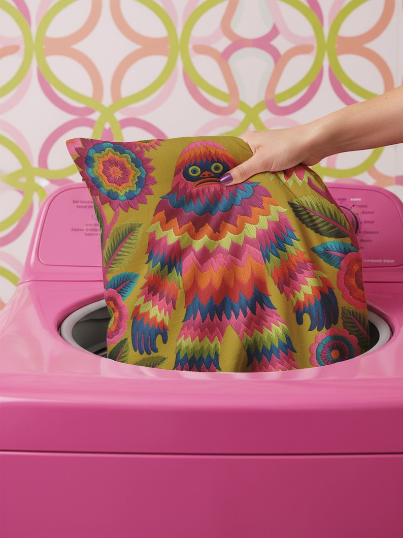 A square faux embroidery pillow featuring a brightly colored maximalist design against a mustard yellow background. In the center stands a stylized, furry Bigfoot or Sasquatch figure rendered in vibrant layers of pink, orange, blue, and lime green with a blue face and yellow eyes. The central creature is surrounded by large, detailed symmetrical pink and blue flowers and leaves. The flat printed fabric creates a highly realistic optical illusion of dimensional crewel embroidery stitches.