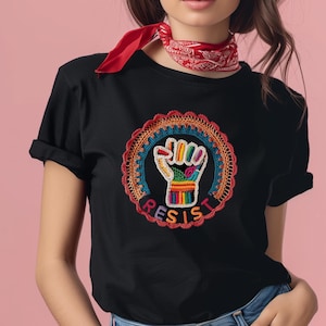Może przedstawiać: Czarny t-shirt z kolorowym haftowanym wzorem uniesionej pięści w okrągłej ramie. Słowo "RESIST" jest wyhaftowane pod pięścią. Koszulka ma krótkie rękawy i okrągły dekolt.