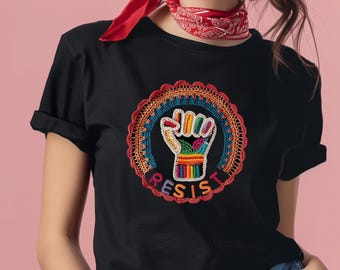 Pode incluir: T-shirt preta com um design bordado colorido de um punho erguido dentro de uma moldura circular. A palavra "RESIST" é bordada abaixo do punho. A camisa tem mangas curtas e gola redonda.