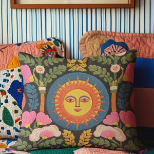 A square folk art sun throw pillow with a dark, charcoal-gray background. The central design is a vibrant celestial sun face in shades of orange, yellow, and red, with defined facial features. The sun is enclosed in a circular blue border with a textured, faux-embroidery pattern. Surrounding the central sun are symmetrical, colorful botanical and floral elements, including pink and white flowers, and green and gold leaves. The overall style is maximalist and bohemian.