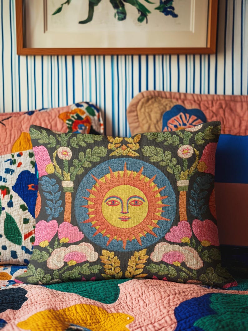 A square folk art sun throw pillow with a dark, charcoal-gray background. The central design is a vibrant celestial sun face in shades of orange, yellow, and red, with defined facial features. The sun is enclosed in a circular blue border with a textured, faux-embroidery pattern. Surrounding the central sun are symmetrical, colorful botanical and floral elements, including pink and white flowers, and green and gold leaves. The overall style is maximalist and bohemian.