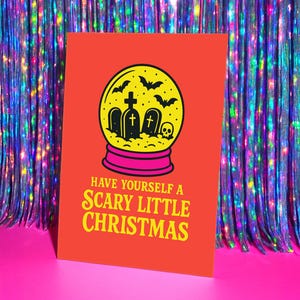 A vibrant, flat-lay photograph of the "Scary Little Christmas" greeting card. The card has a bright orange background with bold, retro yellow text. The central graphic is a snow globe with a yellow glass dome and a hot pink base. Inside the globe is a black silhouette of a spooky graveyard scene, featuring tombstones with crosses, a skull, and a swarm of flying bats. The card is made from premium 120lb silk cover stock with a slight sheen and measures 5x7 inches.