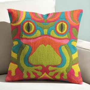 May include: A square cushion with an embroidered frog design in bright colours. The cushion features a vibrant palette of pink, orange, green, yellow, and blue. The frog's eyes are black, and the design is centered on the cushion.
