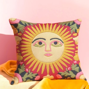 Peut inclure: Coussin carré décoratif avec un motif de visage de soleil brodé. Le soleil a un visage et des rayons jaunes, avec des accents floraux roses et verts sur un fond rouge. Le coussin est sur une surface jaune.
