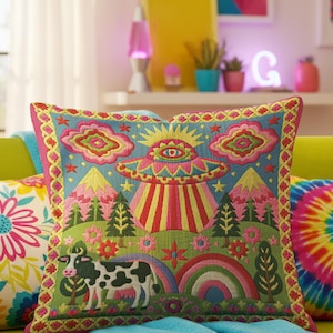 Description of a square throw pillow with a vibrant maximalist alien design. A central UFO with a large all-seeing eye hovers above a landscape with two rainbows, pink mountains, and green pine trees. A black and white spotted cow stands in the foreground. The design is a high-contrast palette of sky blue, magenta, hot pink, and meadow green. The pillow has a faux-embroidery 2D print finish on linen-feel fabric, framed by a yellow and pink scalloped floral border.