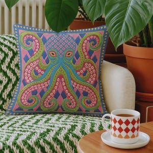A square decorative throw pillow cover featuring a highly symmetrical bright octopus design. The octopus has a turquoise blue body with a diamond patterned head and eight curled tentacles detailed in bright pink and lime green. The background is a repeating diamond argyle pattern in royal blue and light pink. The artwork is a flat two dimensional print that mimics the textured look of intricate crewelwork embroidery. The pillow features a hidden zipper on the bottom edge. No insert included.