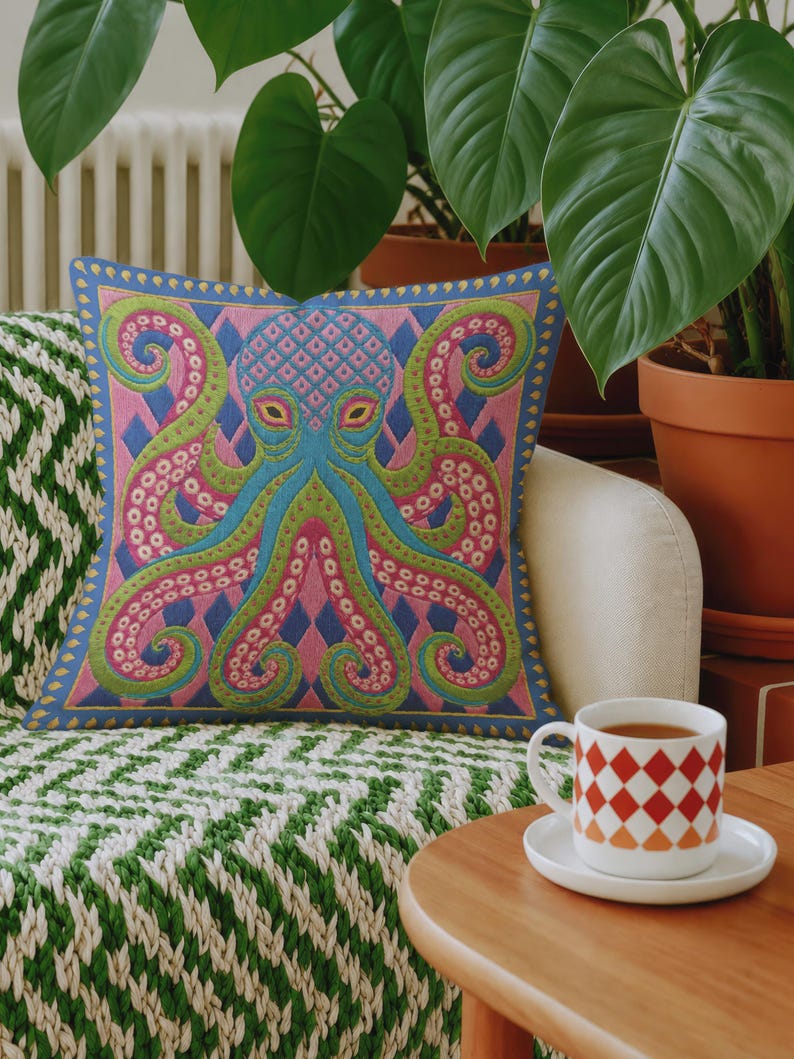 A square decorative throw pillow cover featuring a highly symmetrical bright octopus design. The octopus has a turquoise blue body with a diamond patterned head and eight curled tentacles detailed in bright pink and lime green. The background is a repeating diamond argyle pattern in royal blue and light pink. The artwork is a flat two dimensional print that mimics the textured look of intricate crewelwork embroidery. The pillow features a hidden zipper on the bottom edge. No insert included.