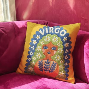 A square throw pillow featuring a vibrant Virgo zodiac design on a golden mustard yellow background. The central illustration shows a terracotta-skinned figure with green hair adorned with white daisies. Blue leaf motifs are printed on the figure’s chest. The word "VIRGO" appears in white bubble letters at the top. The entire design is framed by a royal blue scalloped border, featuring a high-fidelity trompe-l'œil print that mimics 3D embroidery.