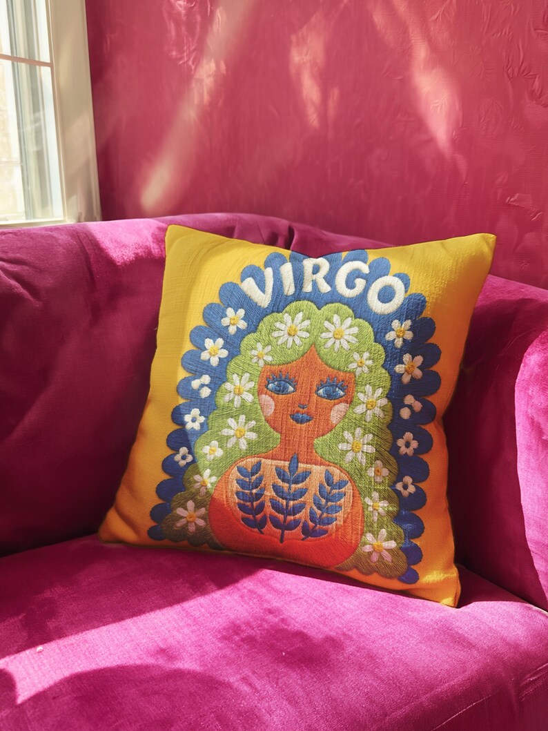 A square throw pillow featuring a vibrant Virgo zodiac design on a golden mustard yellow background. The central illustration shows a terracotta-skinned figure with green hair adorned with white daisies. Blue leaf motifs are printed on the figure’s chest. The word "VIRGO" appears in white bubble letters at the top. The entire design is framed by a royal blue scalloped border, featuring a high-fidelity trompe-l'œil print that mimics 3D embroidery.