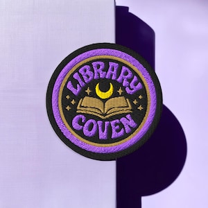 May include: Embroidered patch with the words "LIBRARY COVEN" in purple and gold, surrounding an open book and crescent moon. The patch is circular with a black border and is set against a light purple background.
