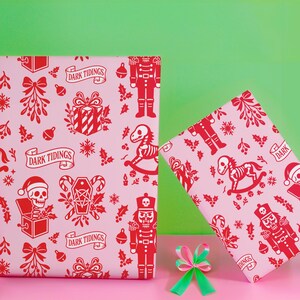 Premium satin finish wrapping paper featuring a repeating pastel goth pattern on a pink background. The design showcases a playful yet spooky aesthetic with high-contrast skeleton nutcrackers, skull jack-in-the-boxes, and rocking horse skeletons in soft pinks, and reds. The paper texture appears semi-glossy. Shown in three available sizes including sheets and rolls, specifically 29 inches wide, suitable for wrapping gifts for holidays, birthdays, or gothic themed occasions.