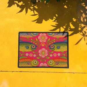 Rectangular indoor outdoor doormat featuring a vibrant hot pink background with a symmetrical maximalist pattern. The design showcases stylized sardines in yellow, lime green, and cobalt blue swimming horizontally towards a central vertical axis of blooming pale pink flowers and green vines. The imagery mimics the texture of vintage needlepoint embroidery but is a flat print on polyester. The mat is framed by a sleek black trim and features a non-slip rubber backing for safety.
