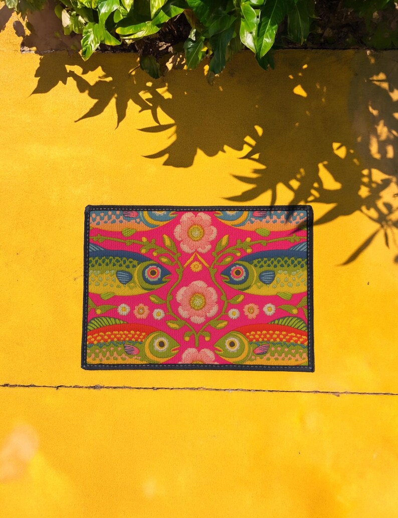 Rectangular indoor outdoor doormat featuring a vibrant hot pink background with a symmetrical maximalist pattern. The design showcases stylized sardines in yellow, lime green, and cobalt blue swimming horizontally towards a central vertical axis of blooming pale pink flowers and green vines. The imagery mimics the texture of vintage needlepoint embroidery but is a flat print on polyester. The mat is framed by a sleek black trim and features a non-slip rubber backing for safety.