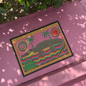 A rectangular doormat featuring a colorful 2D flat print designed to mimic raised, textured yarn embroidery. The central graphic showcases a large dark grey and yellow alligator resting in wavy water stripes of red, bright blue, and yellow. The background is hot pink, decorated with two green palm trees, a leafy green agave plant, and two stylized suns. The entire design is enclosed by a yellow border with repeating circle motifs and finished with a sleek black outer edge border trim.