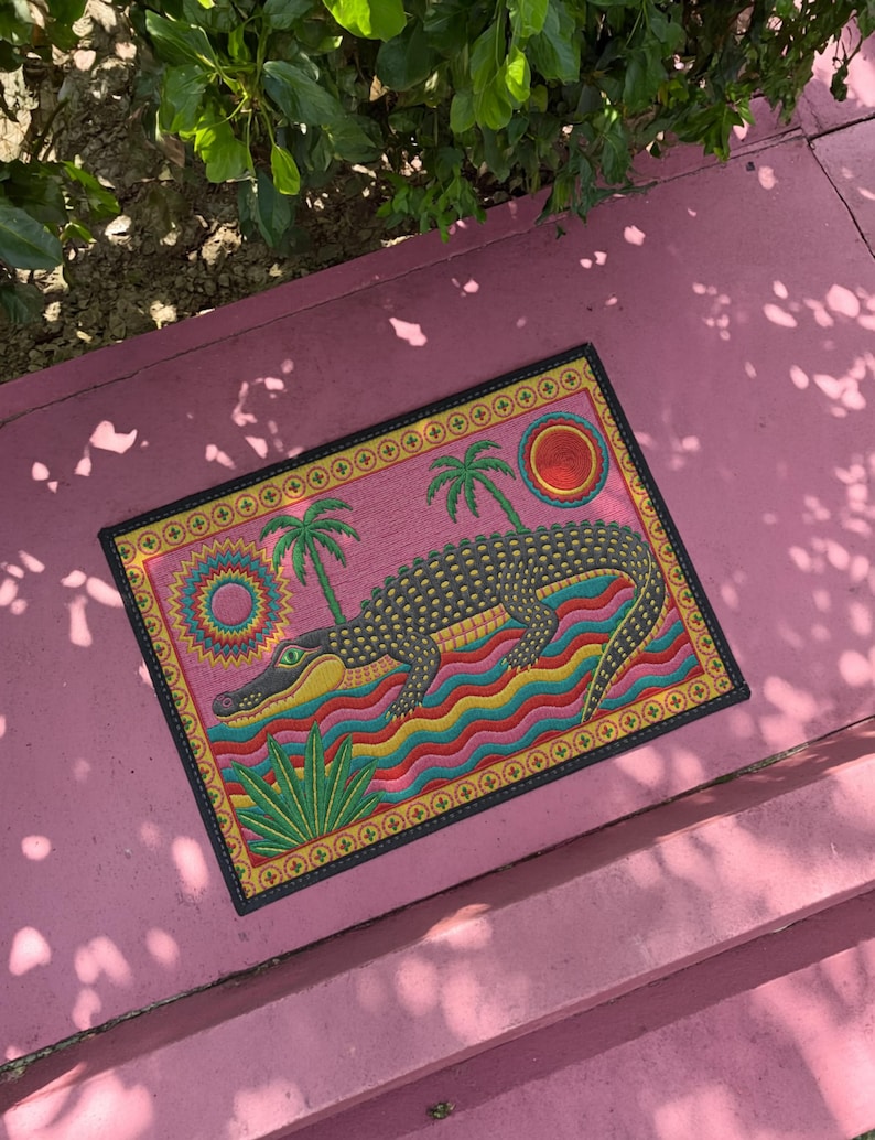 A rectangular doormat featuring a colorful 2D flat print designed to mimic raised, textured yarn embroidery. The central graphic showcases a large dark grey and yellow alligator resting in wavy water stripes of red, bright blue, and yellow. The background is hot pink, decorated with two green palm trees, a leafy green agave plant, and two stylized suns. The entire design is enclosed by a yellow border with repeating circle motifs and finished with a sleek black outer edge border trim.