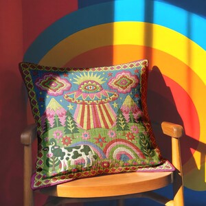 Description of a square throw pillow with a vibrant maximalist alien design. A central UFO with a large all-seeing eye hovers above a landscape with two rainbows, pink mountains, and green pine trees. A black and white spotted cow stands in the foreground. The design is a high-contrast palette of sky blue, magenta, hot pink, and meadow green. The pillow has a faux-embroidery 2D print finish on linen-feel fabric, framed by a yellow and pink scalloped floral border.