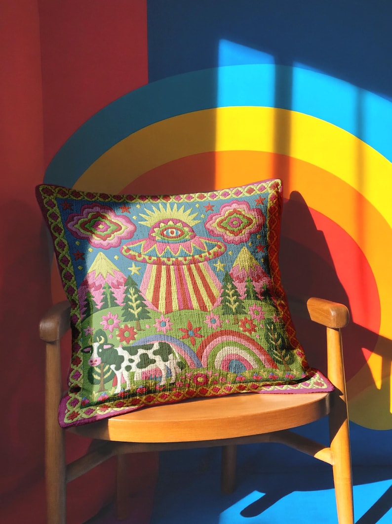 Description of a square throw pillow with a vibrant maximalist alien design. A central UFO with a large all-seeing eye hovers above a landscape with two rainbows, pink mountains, and green pine trees. A black and white spotted cow stands in the foreground. The design is a high-contrast palette of sky blue, magenta, hot pink, and meadow green. The pillow has a faux-embroidery 2D print finish on linen-feel fabric, framed by a yellow and pink scalloped floral border.