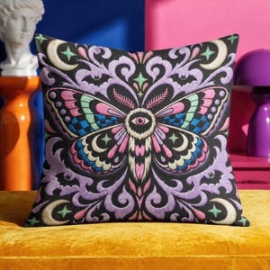May include: A square throw pillow featuring a black background with a central moth illustration. The moth has pink, blue, and green wings with a single eye in the center. The design includes purple swirling patterns, crescent moons, and stars.