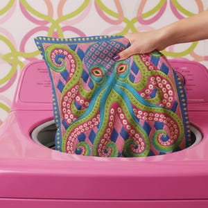 A square decorative throw pillow cover featuring a highly symmetrical bright octopus design. The octopus has a turquoise blue body with a diamond patterned head and eight curled tentacles detailed in bright pink and lime green. The background is a repeating diamond argyle pattern in royal blue and light pink. The artwork is a flat two dimensional print that mimics the textured look of intricate crewelwork embroidery. The pillow features a hidden zipper on the bottom edge. No insert included.