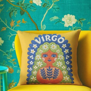 A square throw pillow featuring a vibrant Virgo zodiac design on a golden mustard yellow background. The central illustration shows a terracotta-skinned figure with green hair adorned with white daisies. Blue leaf motifs are printed on the figure’s chest. The word "VIRGO" appears in white bubble letters at the top. The entire design is framed by a royal blue scalloped border, featuring a high-fidelity trompe-l'œil print that mimics 3D embroidery.