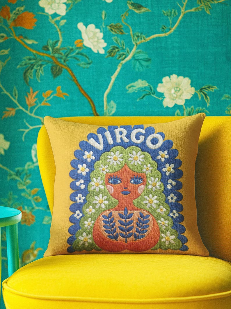 A square throw pillow featuring a vibrant Virgo zodiac design on a golden mustard yellow background. The central illustration shows a terracotta-skinned figure with green hair adorned with white daisies. Blue leaf motifs are printed on the figure’s chest. The word "VIRGO" appears in white bubble letters at the top. The entire design is framed by a royal blue scalloped border, featuring a high-fidelity trompe-l'œil print that mimics 3D embroidery.