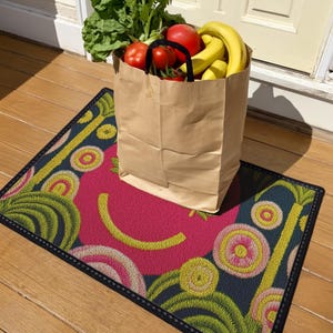 A rectangular doormat featuring a large hot pink smiley face on a deep teal background. The smiley face has green cannabis leaves for eyes and a yellow stitched-style smile. The central face is surrounded by stylized green foliage arches and concentric circles in pink, yellow, and white, resembling abstract flowers. The design mimics a vintage needlepoint or tapestry texture but is a flat print. The mat has a solid black trim border.
