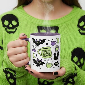White ceramic mug with vibrant purple interior and matching C-handle. Features a full wrap-around, 'spooky cute' pastel goth design. Central graphic is a lime green coffee cup with 'CERTIFIED NIGHT CREATURE' in black text. Surrounding illustrations include grumpy black bats, a steaming green coffee cup, a purple mug, vampire fangs, crescent moon, black cherries, and an alarm clock. Design uses bold graphics in black, lime green, and purple, accented with small black stars. Pastel Goth vibes.