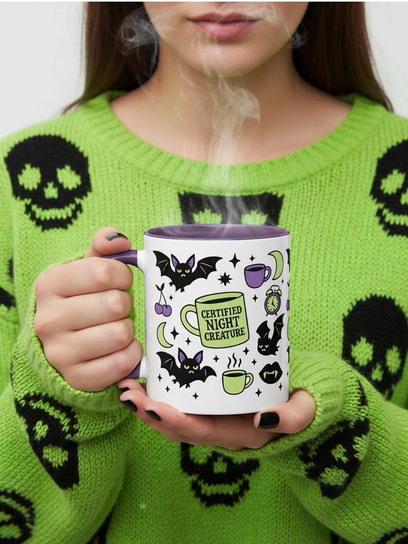 White ceramic mug with vibrant purple interior and matching C-handle. Features a full wrap-around, 'spooky cute' pastel goth design. Central graphic is a lime green coffee cup with 'CERTIFIED NIGHT CREATURE' in black text. Surrounding illustrations include grumpy black bats, a steaming green coffee cup, a purple mug, vampire fangs, crescent moon, black cherries, and an alarm clock. Design uses bold graphics in black, lime green, and purple, accented with small black stars. Pastel Goth vibes.
