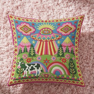 Description of a square throw pillow with a vibrant maximalist alien design. A central UFO with a large all-seeing eye hovers above a landscape with two rainbows, pink mountains, and green pine trees. A black and white spotted cow stands in the foreground. The design is a high-contrast palette of sky blue, magenta, hot pink, and meadow green. The pillow has a faux-embroidery 2D print finish on linen-feel fabric, framed by a yellow and pink scalloped floral border.