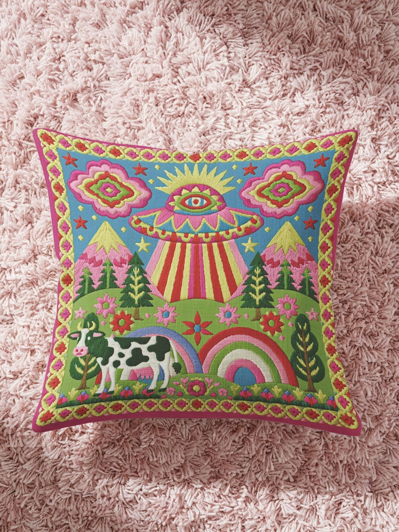 Description of a square throw pillow with a vibrant maximalist alien design. A central UFO with a large all-seeing eye hovers above a landscape with two rainbows, pink mountains, and green pine trees. A black and white spotted cow stands in the foreground. The design is a high-contrast palette of sky blue, magenta, hot pink, and meadow green. The pillow has a faux-embroidery 2D print finish on linen-feel fabric, framed by a yellow and pink scalloped floral border.
