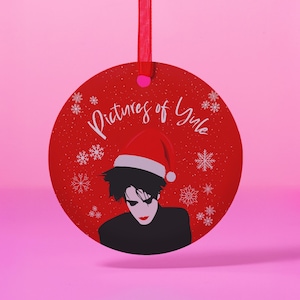 May include: A red round Christmas ornament with a white snowflake pattern. The ornament features a black and white illustration of a person wearing a red Santa hat. The text "Pictures of Yule" is written in white on the ornament.