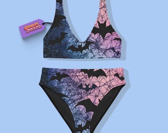Pode incluir: Um conjunto de biquíni de duas peças com top de decote em V profundo e calcinha de cintura alta. O tecido apresenta um padrão degradê de azul para rosa, sobreposto com silhuetas de morcegos pretos e uma estampa de teia. Uma etiqueta roxa diz Honey Dazed.