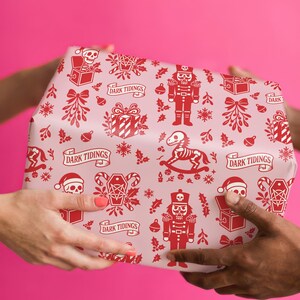 Premium satin finish wrapping paper featuring a repeating pastel goth pattern on a pink background. The design showcases a playful yet spooky aesthetic with high-contrast skeleton nutcrackers, skull jack-in-the-boxes, and rocking horse skeletons in soft pinks, and reds. The paper texture appears semi-glossy. Shown in three available sizes including sheets and rolls, specifically 29 inches wide, suitable for wrapping gifts for holidays, birthdays, or gothic themed occasions.
