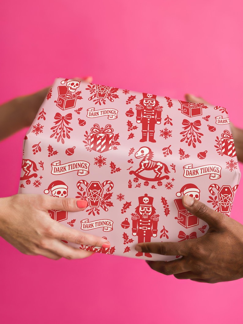 Premium satin finish wrapping paper featuring a repeating pastel goth pattern on a pink background. The design showcases a playful yet spooky aesthetic with high-contrast skeleton nutcrackers, skull jack-in-the-boxes, and rocking horse skeletons in soft pinks, and reds. The paper texture appears semi-glossy. Shown in three available sizes including sheets and rolls, specifically 29 inches wide, suitable for wrapping gifts for holidays, birthdays, or gothic themed occasions.