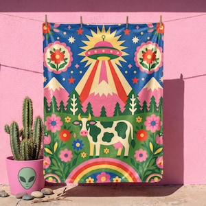 May include: A rectangular blanket featuring a colorful graphic print of a cow standing in a field with flowers, pine trees, and mountains. Above the landscape, a yellow sunburst surrounds a flying saucer emitting a beam of light and red stars.