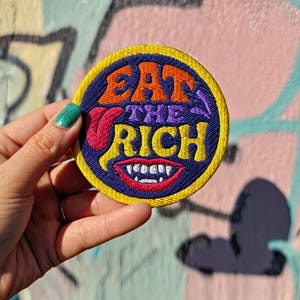 Peut inclure: Écusson brodé rond avec l'inscription "EAT THE RICH" en orange, violet et rouge, avec un motif de bouche souriante. L'écusson a une bordure jaune et un fond bleu.