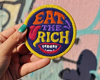 May include: Embroidered patch with the words "EAT THE RICH" in orange, purple, and red, with a smiling mouth graphic. The patch has a yellow border and a blue background. The patch is round.