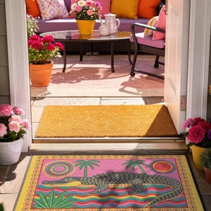 A rectangular doormat featuring a colorful 2D flat print designed to mimic raised, textured yarn embroidery. The central graphic showcases a large dark grey and yellow alligator resting in wavy water stripes of red, bright blue, and yellow. The background is hot pink, decorated with two green palm trees, a leafy green agave plant, and two stylized suns. The entire design is enclosed by a yellow border with repeating circle motifs and finished with a sleek black outer edge border trim.