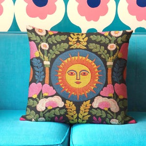 A square folk art sun throw pillow with a dark, charcoal-gray background. The central design is a vibrant celestial sun face in shades of orange, yellow, and red, with defined facial features. The sun is enclosed in a circular blue border with a textured, faux-embroidery pattern. Surrounding the central sun are symmetrical, colorful botanical and floral elements, including pink and white flowers, and green and gold leaves. The overall style is maximalist and bohemian.