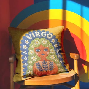 A square throw pillow featuring a vibrant Virgo zodiac design on a golden mustard yellow background. The central illustration shows a terracotta-skinned figure with green hair adorned with white daisies. Blue leaf motifs are printed on the figure’s chest. The word "VIRGO" appears in white bubble letters at the top. The entire design is framed by a royal blue scalloped border, featuring a high-fidelity trompe-l'œil print that mimics 3D embroidery.